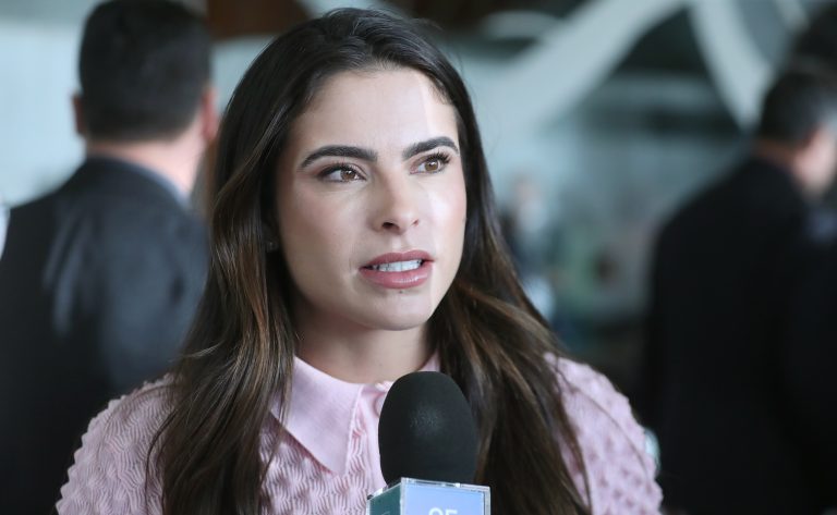 Deputada Yandra Moura fala ao microfone. Ela é branca, tem o cabelo longo e escuro e usa uma blusa rosa